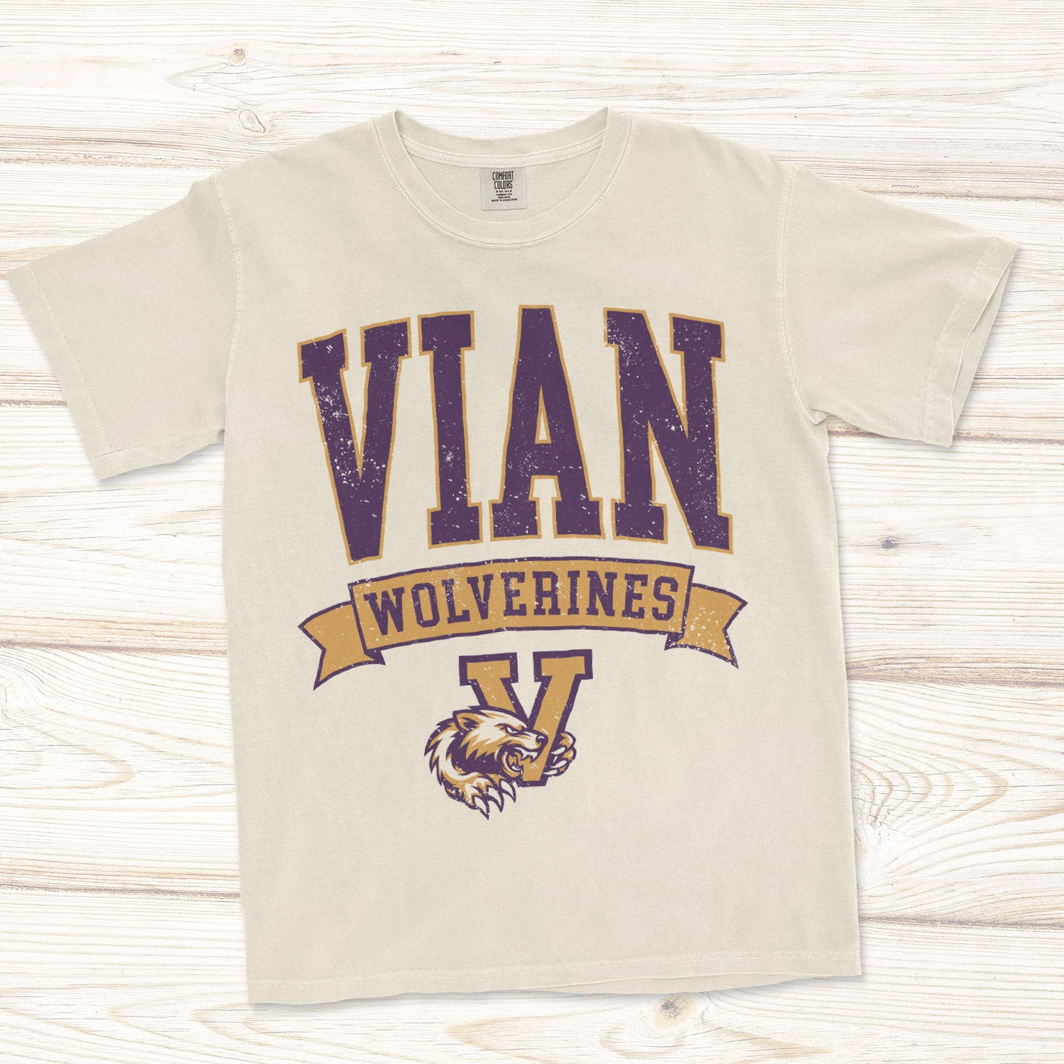 A cream-colored short-sleeve t-shirt with a distressed graphic reading 'VIAN WOLVERINES' in purple and gold, featuring a wolverine mascot with his claw wrapped around the Vian victory V logo.